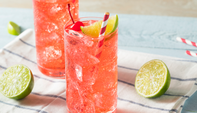 Citric Acid 500 (Cherry limeade) in a tall clear glass with a straw, cherry, and lime wedge. The glass is on a white and blue striped fabric with limes halves and straws scattered around