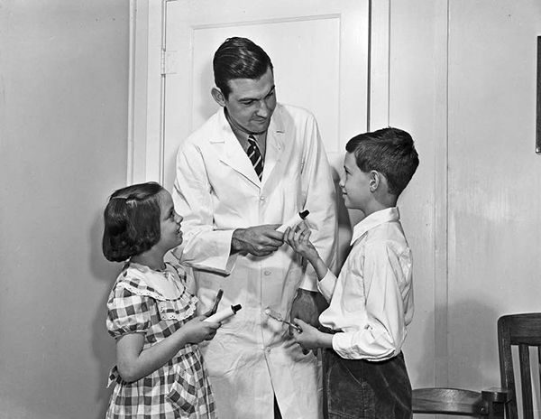 A dentist in a lab coat gives small toothpaste tubes to two children