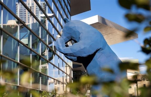 Photo of the Colorado Convention Center, glass windows on the left and the big blue bear statue in the center surrounded by vegetation
