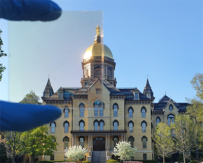 A hand holds a transparent film in front of a view of a building, demonstrating that the film doesn’t block visible light.