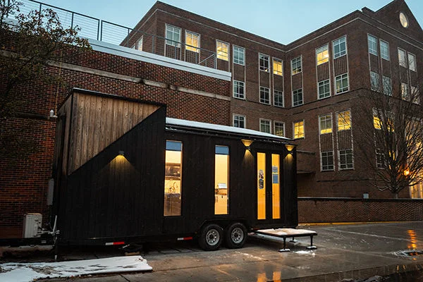 A trailer-sized tiny house on wheels sits outside a multi-story academic building.