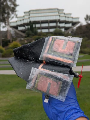 Outside in front of a university building, a gloved hand holds a small, black glider that has two rectangles of copper metal covered by a plastic film on the back of the wings.