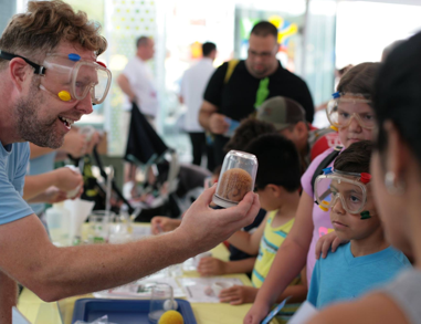 Activity facilitator holds up a jar for a group of kids wearing safety goggles to examine