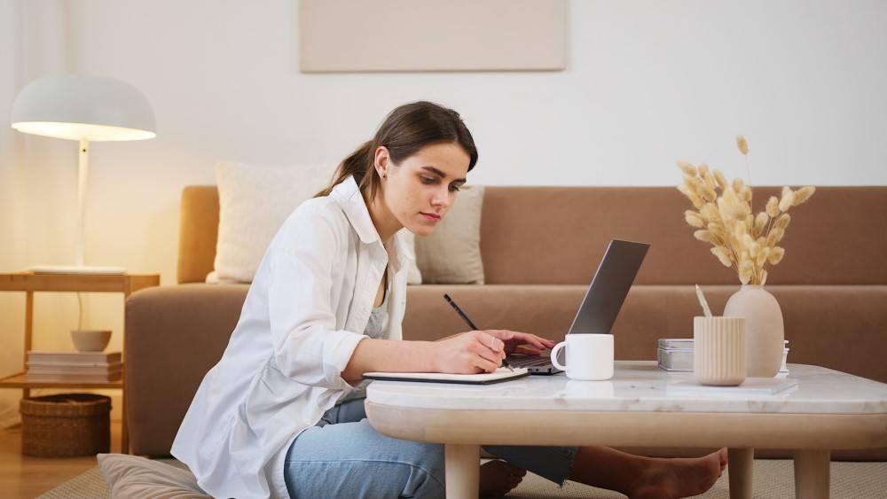 Woman sitting on floor writing in notebook with laptop 