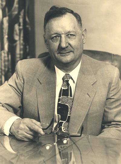 Sepia photo of Harley Wilhelm. He is wearing a suit and seated at a desk, holding a pair of glasses in his right hand.