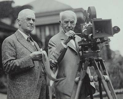 Black and white photograph of George Eastman and Thomas Edison, both wearing suits standing behind a motion picture camera