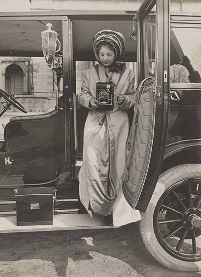 The "Kodak girl" stands in the door of a car from the 1910s, holding a Kodak camera