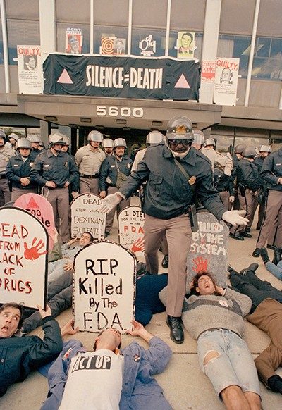 Demonstrators lie on the ground in front of the FDA headquarters, holding signs in the shape of graves. A line of law enforcement officers stand between them and the building.