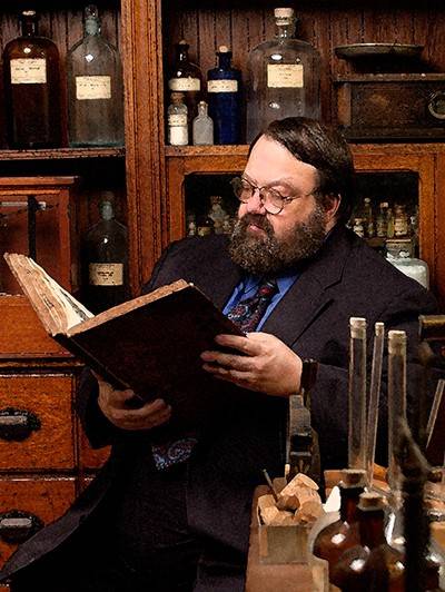 Jensen, wearing a suit, reads a large, old book while seated in the museum's reproduction of a chemical laboratory