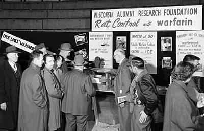 A sign reads "Rat control and warfarin" and a group of people investigate the display