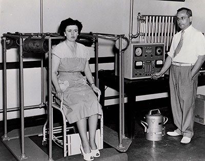 A woman sitting in chair with a measurement device behind her head on left, a man standing next to a control panel for the device on the right.