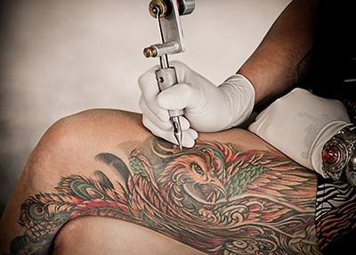 A gloved hand holding a tattoo machine working on a piece on another person's leg