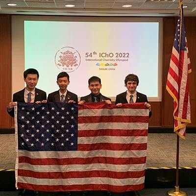 Four high school students wearing suits, holding an American flag in front of them