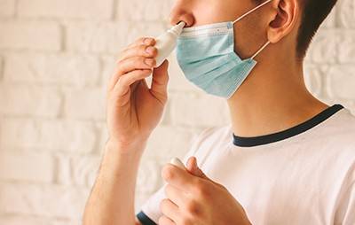 A man wearing a mask holds a bottle of nasal spray to his nose.