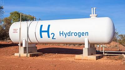 A large white tank with H2 and Hydrogen written on the side in blue sits horizontally on concrete blocks in front of a mound of reddish soil 
