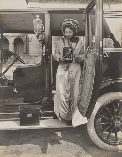 A woman holding a Kodak camera sitting in car with an open door.