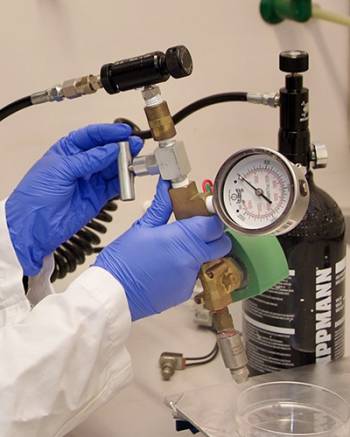 A pair of gloved hands hold the MOF-Jet device, composed of metal fittings attached together, a pressure gauge, a tank of compressed gas and a firing tip.