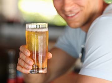 A man with a slight smile leaning over a bar, holding a tall clear glass of lager