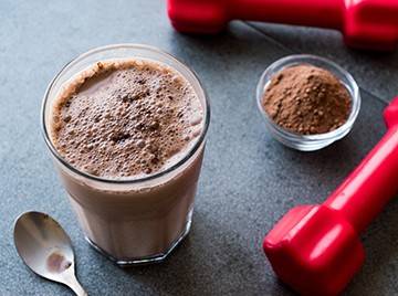 Glass cup with cocoa mixed into the liquid, with a metal spoon, a pinch bowl of powdered cocoa, and two red hand weights on the surface nearby