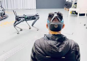 A person wears an augmented reality headset and electrical sensors, controlling a four-legged robot standing away from them.