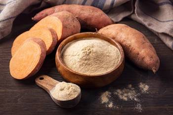 A bowl and spoon filled with sweet potato flour, surrounded by whole and sliced sweet potatoes.