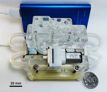 Clear plastic pieces are stacked and connected by tubing to make a microfluidic chip. A blue portable battery pack stands upright behind the microfluidic chip. A 20-millimeter-wide bar provides scale for the device.