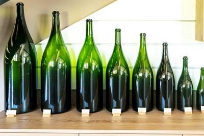 Eight green wine bottles of decreasing sizes from left to right are lined up on a wooden table.