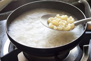 A metal spoon holds cooked elbow macaroni above a pot of boiling water with pasta in it