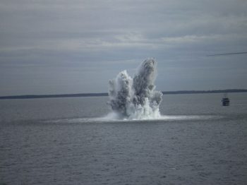 A gray and white geyser of water erupting from a calm, flat ocean surface