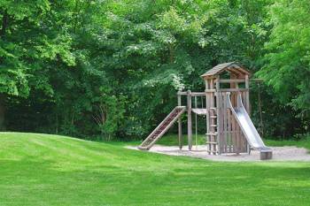 A wooden playground and sandy area sitting in a bright green park.