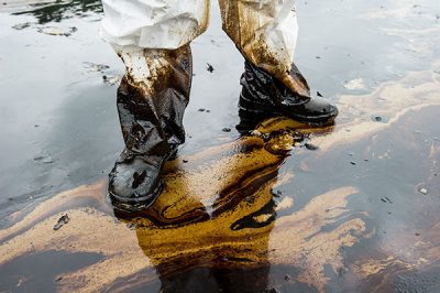 A person standing on spilled, crude oil on a beach, with oil covering their shoes and pants.