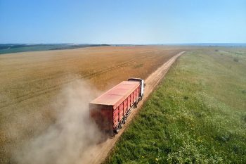 A cargo truck driving on a dirt road through farm fields, with a dust cloud kicked up behind it.