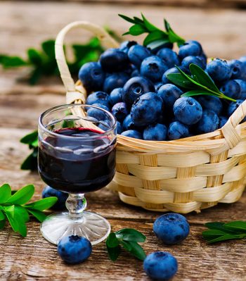 A glass of blueberry wine sits in front of a basket of blueberries on a wooden table.